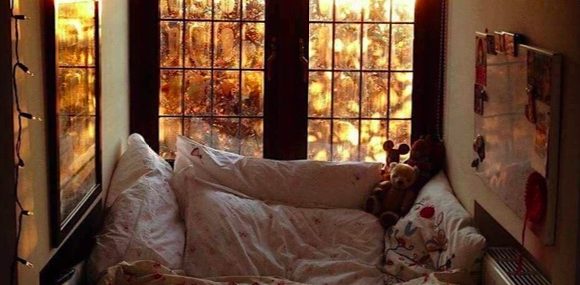 A bunch of fluffy pillows and a stuffed animal on a chair. Behind it is a picture window showing golden leaved trees, in fall.