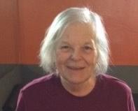 A headshot of Jean Riseman. She is looking directly at the camera and smiling. She has shoulder length, straight white hair and is wearing a burgundy top.