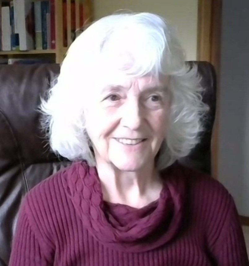 Portrait of Alison Miller. An elder with shoulder length white, curly hair, smiling and wearing a burgundy sweater.