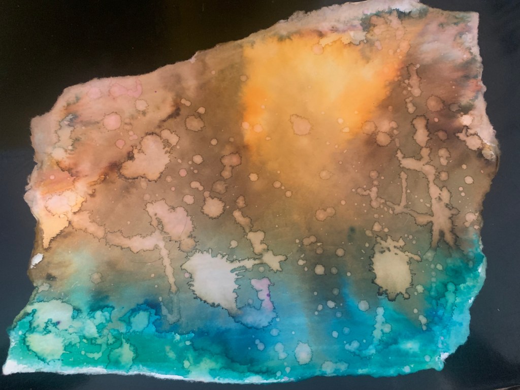 An irregularly shaped piece of paper with torn edges. It's filled with areas of blended blue, green, brown, orange, yellow, and gold in abstract shapes. It also has various water marks on it, creating a kind of splatter design.