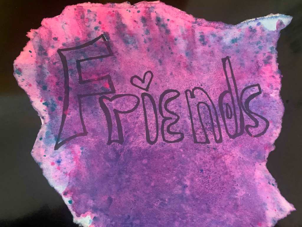 A curled piece of paper dyed in mottled pink, purple, and blue, with the word "Friends" in the center. The word "Friends" has a heart as the dot of the i.