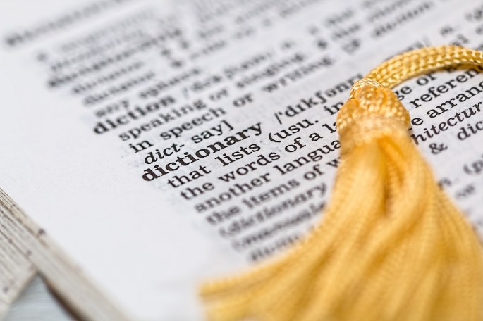 An open page of a dictionary with a gold tassel from a bookmark laying over it. It's open to the word "dictionary" and suggests someone has been looking in the dictionary for a definition of that word.