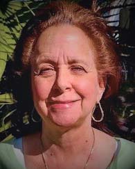 Portrait of Ellen Lacter. An older woman with copper colored hair, large hoop earrings, and a green shirt, looking directly at the camera and smiling.