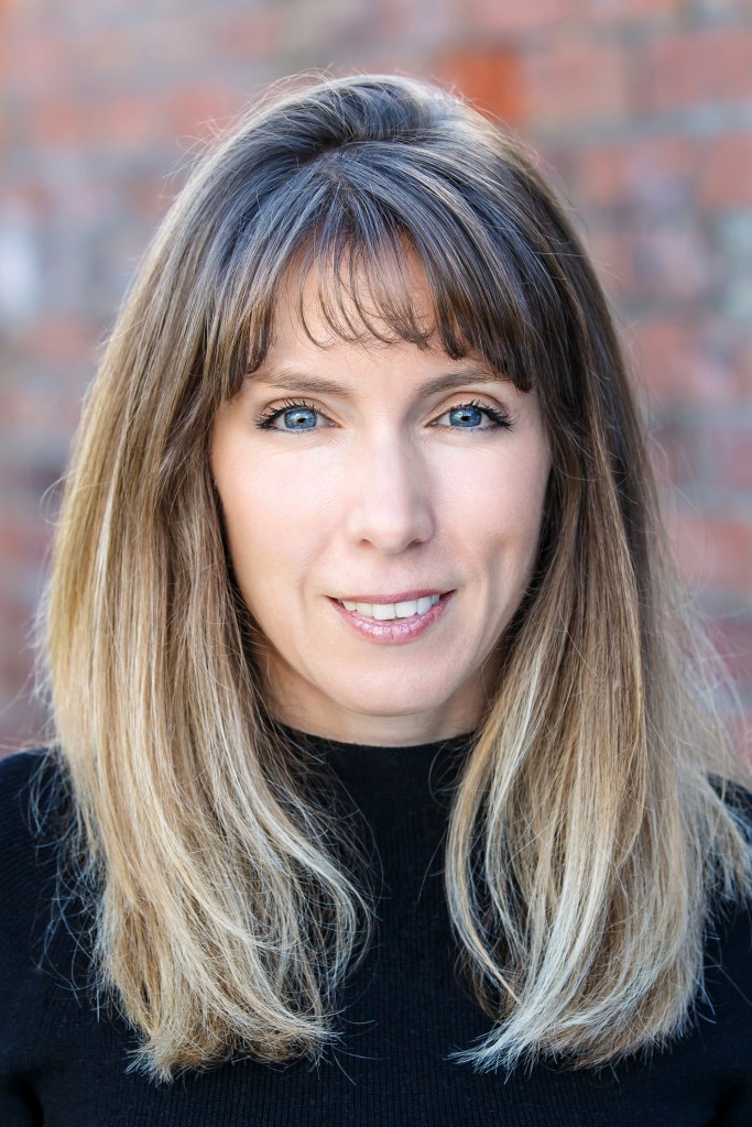 A headshot of Elly Hanson - a woman with straight, long brown hair that's lightened near the ends. She's smiling at the camera and wearing a black sweater.