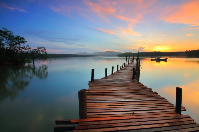A wooden pier stretching out into the distance over a calm lake. The sun is just above the horizon and while the sky is largely blue, some nearby streaks of cloud are colored orange. The image has a feeling of purpose (heading in a specific direction), peacefulness, and potential.